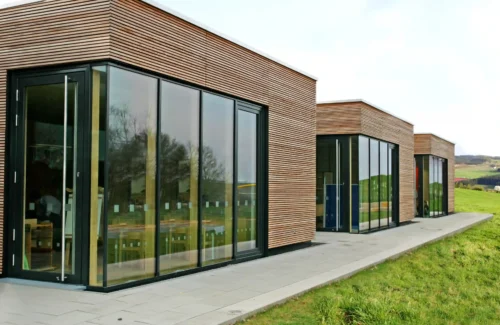 Moderne Holzhäuser auf einer grünen Wiese mit mit großen Holz-Aluminium-Fenstern.